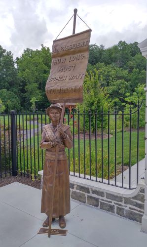 Suffragist statue in front of replica white house fence with sign reading how long must women wait for liberty